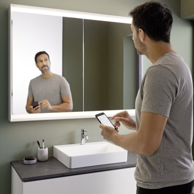 Moški nadzoruje osvetlitev omarice z ogledalom Geberit ONE s svojim pametnim telefonom (© Geberit)